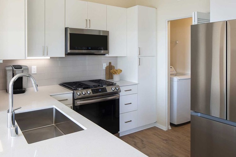 Renovated Package I kitchen featuring white cabinetry, white quartz countertops, stainless steel appliances, and hard surface flooring. In-unit laundry with full sized washer and dryer