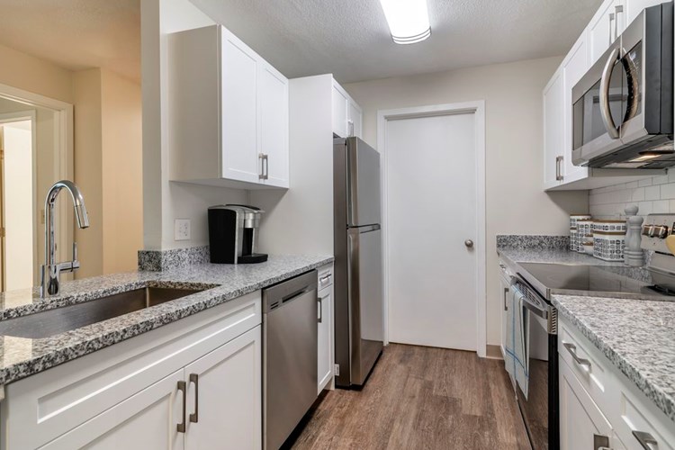 Renovated Package I kitchen with stainless steel appliances, grey speckled granite countertops, white cabinetry, white tile backsplash, and hard surface flooring
