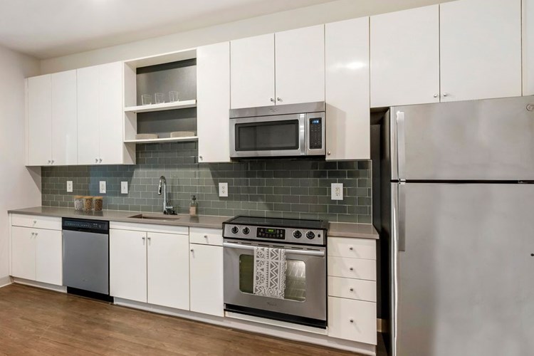 Renovated Package I kitchen with stainless steel appliances, grey quartz countertops, white cabinetry, grey tile backsplash, and hard surface flooring