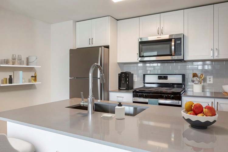 Newly renovated Finish Package II kitchen with white cabinetry, grey quartz countertops, extended hard surface flooring in select units, stainless steel appliances, and tile backsplash