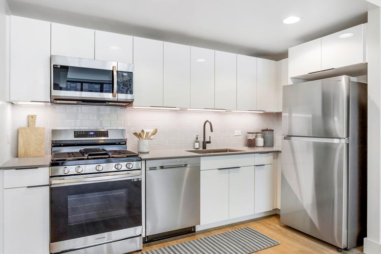 Renovated Package I kitchen with grey quartz countertops, white cabinetry, light grey tile backsplash, stainless steel appliances, and hard surface flooring