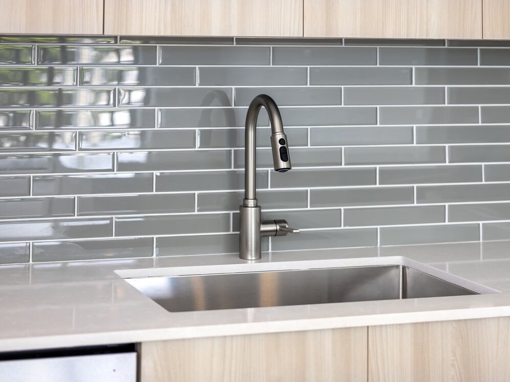 Upgrade Collection I Three-bedroom apartment kitchen with light wood cabinetry, grey tile backsplash, white quartz countertop, and stainless steel appliances