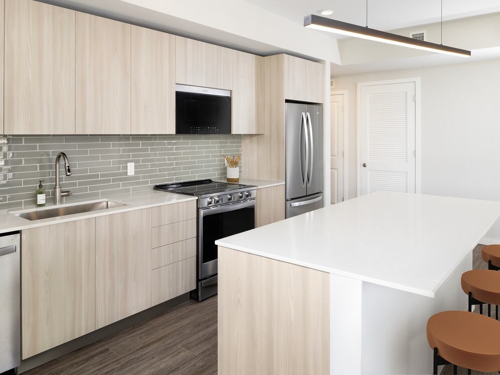 Upgrade Collection I Three-bedroom apartment kitchen with light wood cabinetry, grey tile backsplash, white quartz countertop, and stainless steel appliances