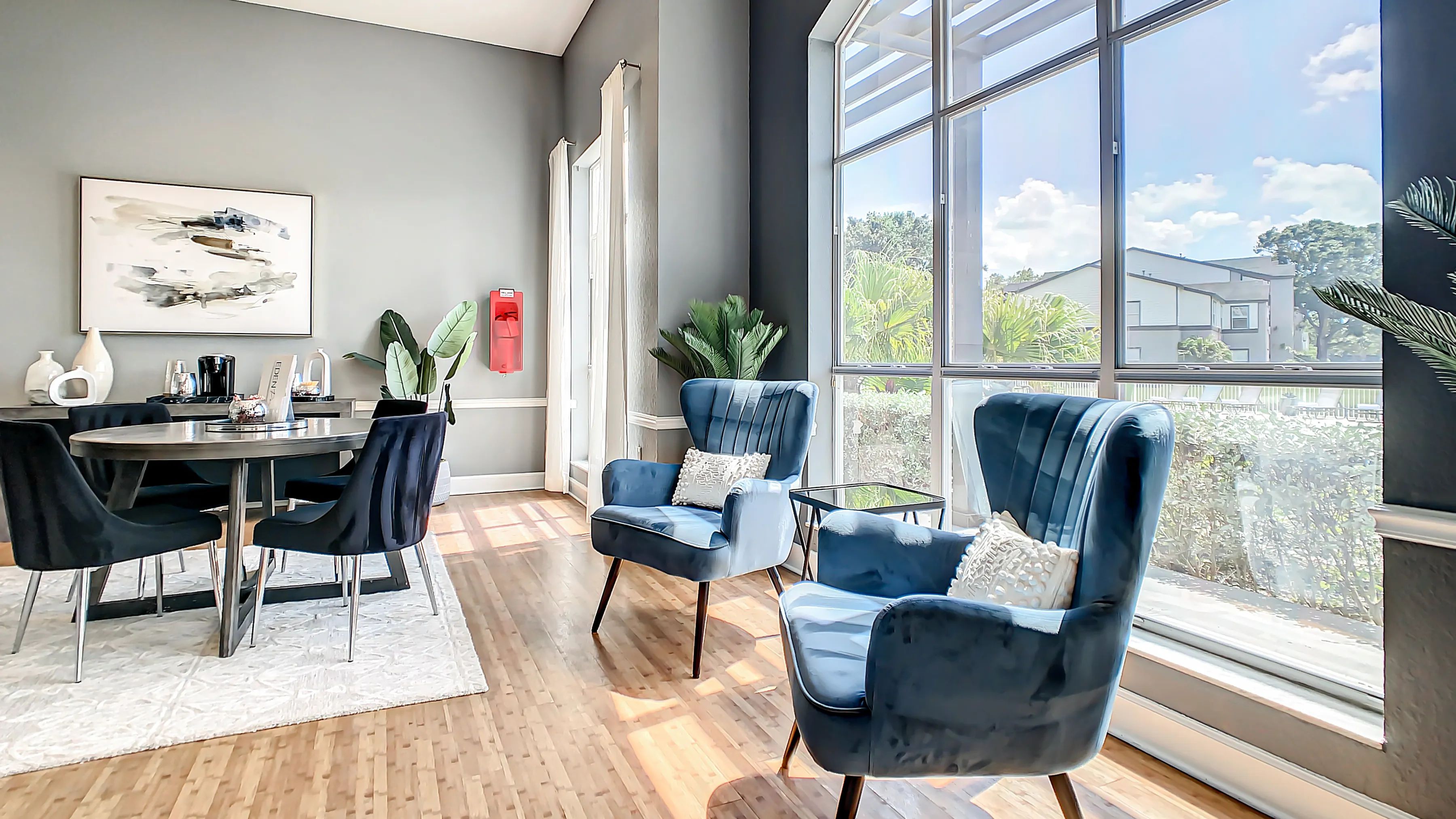 A corner of the leasing office, featuring tables with plush velvet chairs and additional velvet armchairs against a large window that is overlooking the pool deck.