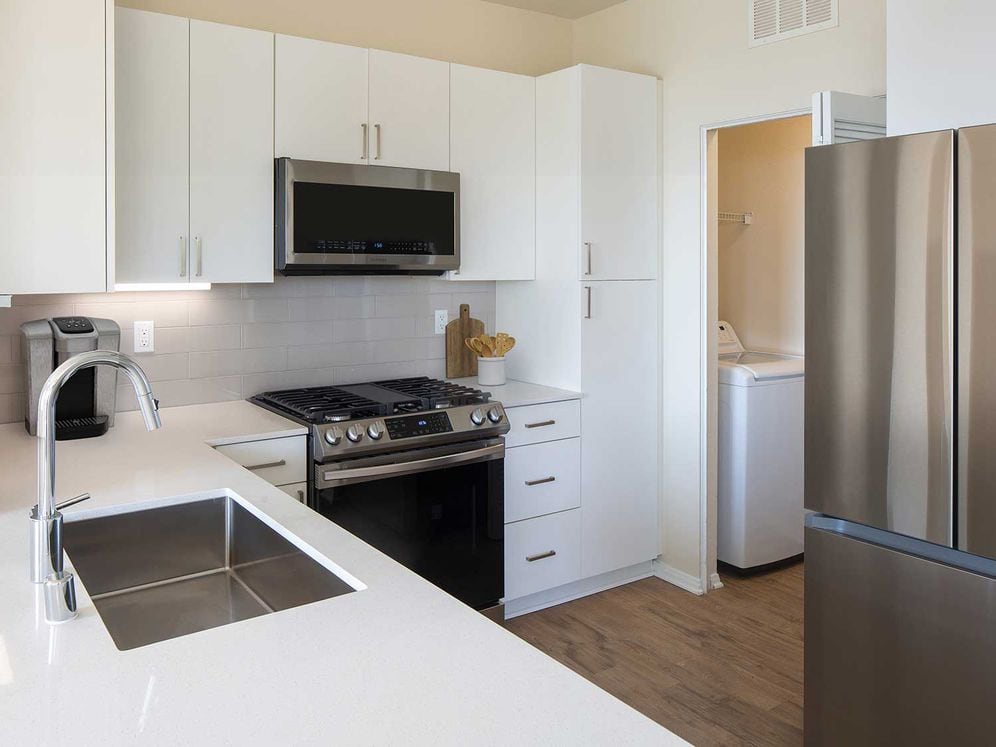 Renovated Package I kitchen featuring white cabinetry, white quartz countertops, stainless steel appliances, and hard surface flooring. In-unit laundry with full sized washer and dryer