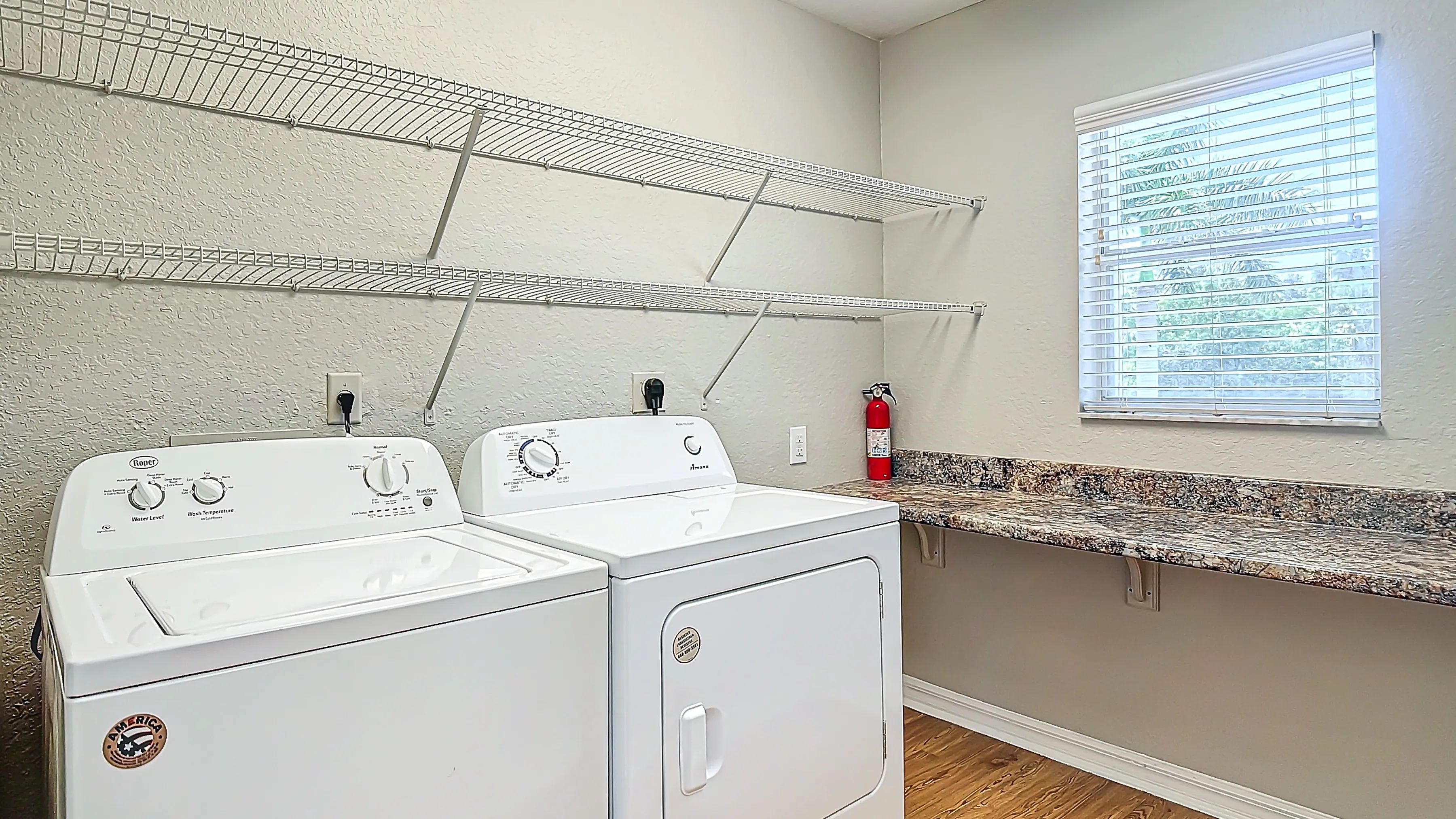 This in-unit laundry room offers ample space for all your laundry needs, featuring a washer, dryer, built-in shelving, and a large countertop beneath a bright window.