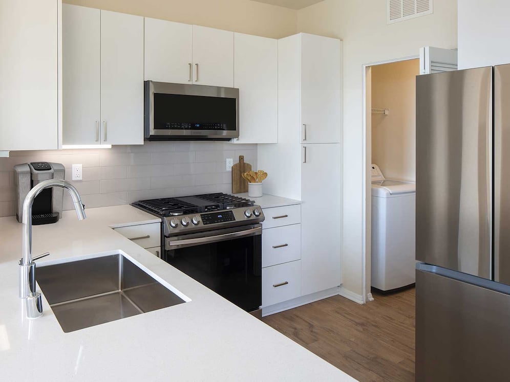 Renovated Package I kitchen featuring white cabinetry, white quartz countertops, stainless steel appliances, and hard surface flooring. In-unit laundry with full sized washer and dryer