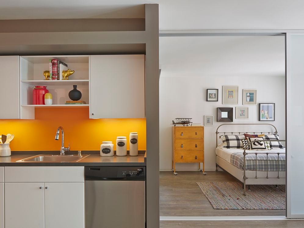 Open concept kitchen with white cabinetry, dark grey countertops with color backsplash stainless steel appliances and bedroom with sliding barn door and hard surface flooring