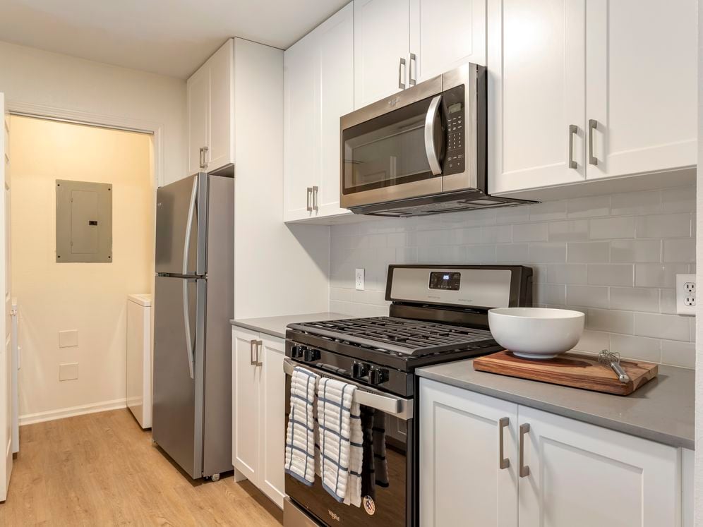 Renovated Package I kitchen with white cabinetry, grey quartz countertops, stainless steel appliances, hard surface flooring, and tile backsplash. In-unit washer and dryer