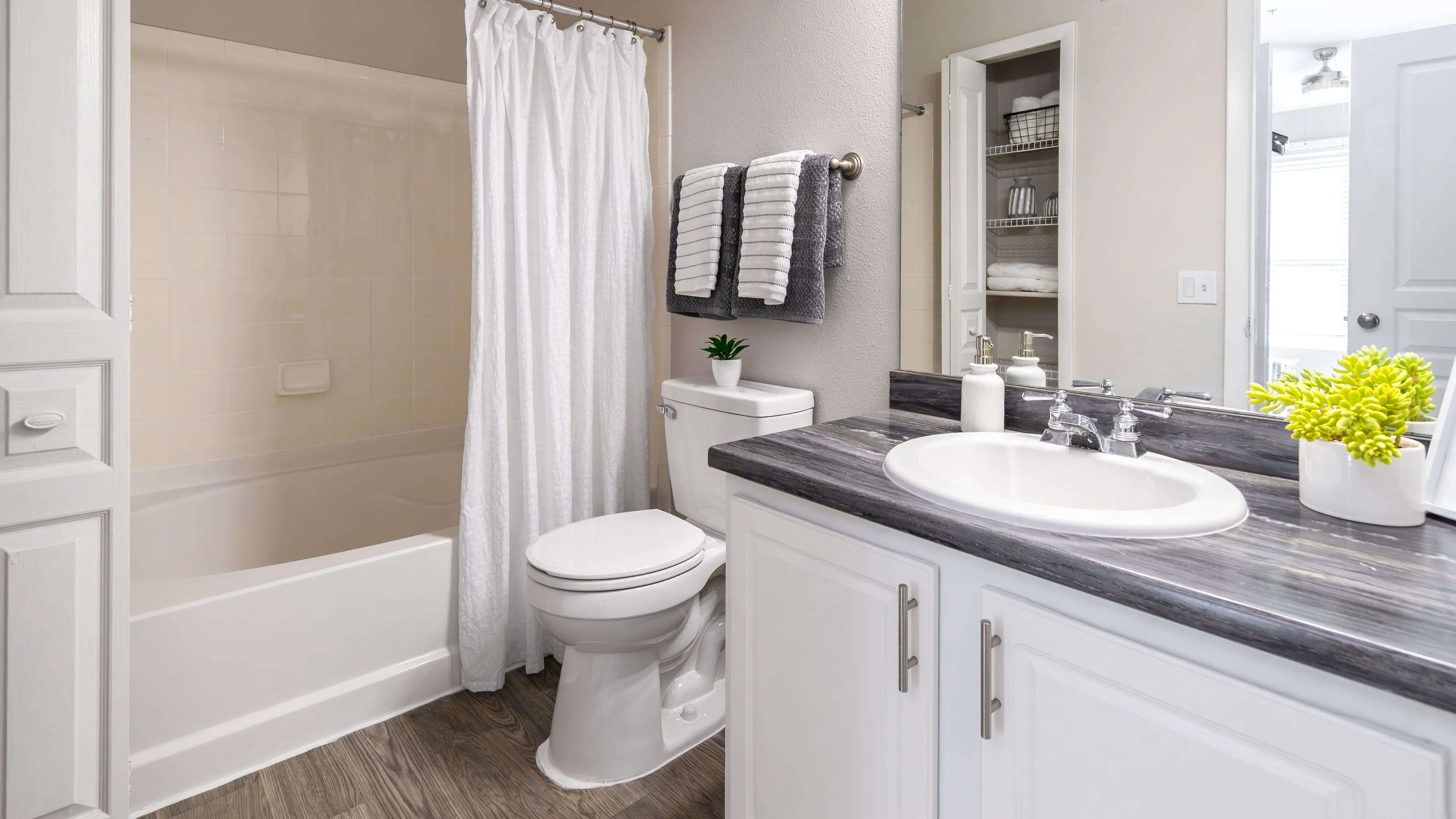 Enjoy a fresh start every day in this stylish bathroom featuring black fusion countertops and white cabinetry, a large mirror and wood-style flooring.