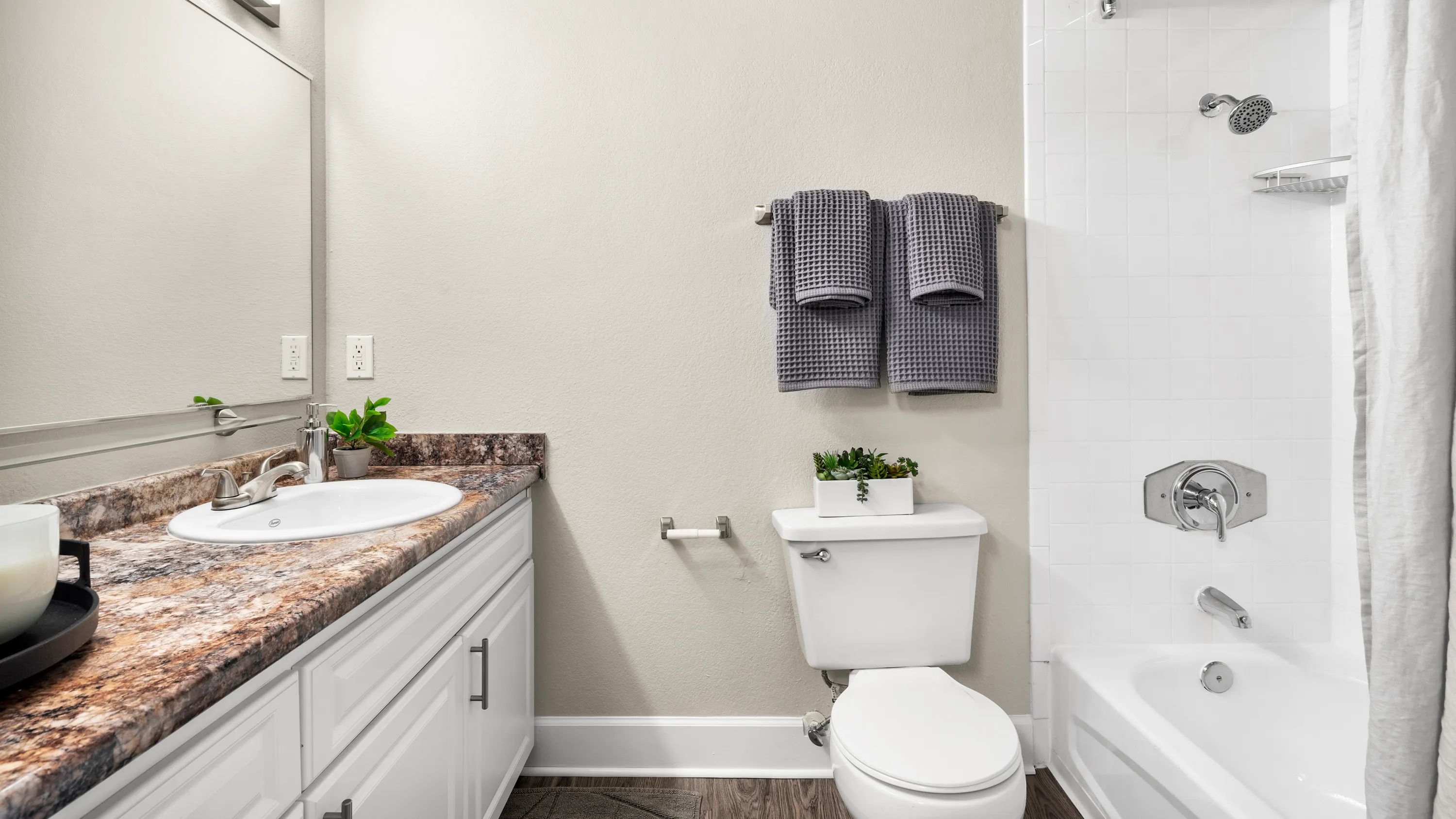 Step into our revitalized bathroom retreats, featuring deluxe granite-style countertops, grand wall-to-wall mirrors, and wood-style flooring with white tile surround for a luxurious and vibrant experience. 