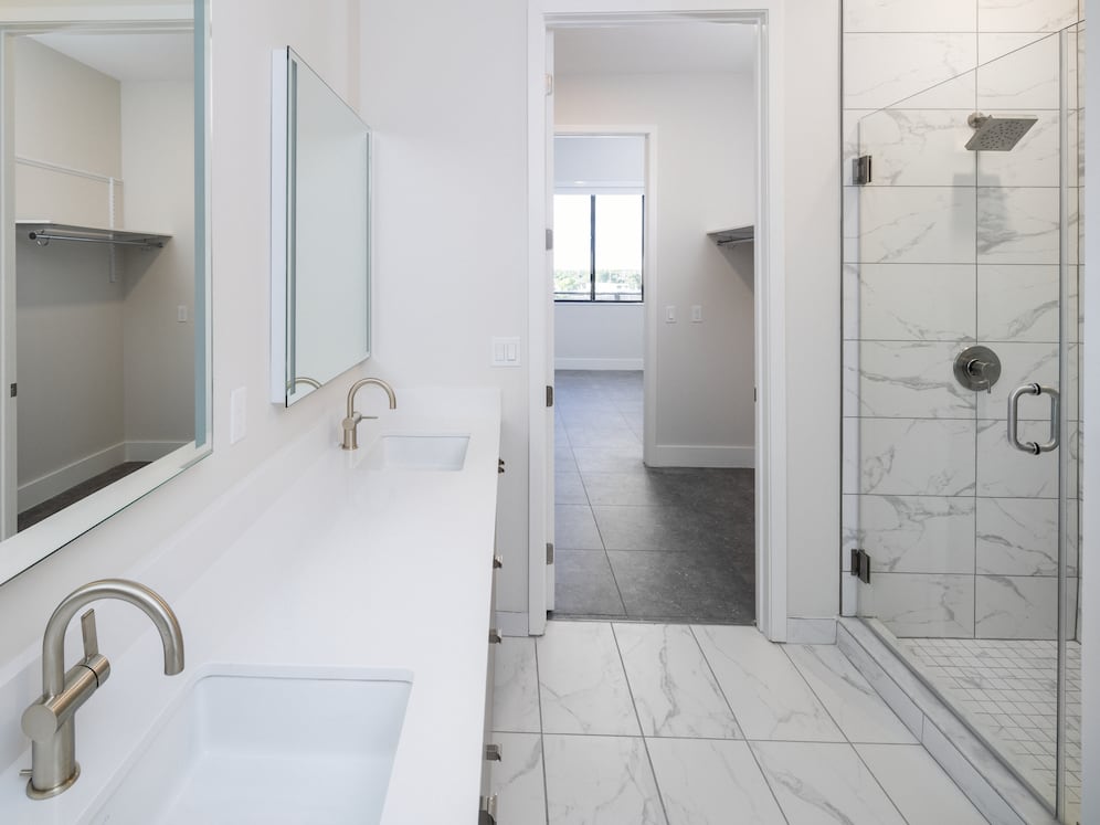 Signature Collection bathroom with grey cabinetry, white quartz countertop, lighted vanity mirrors, porcelain shower surround, and porcelain tile flooring