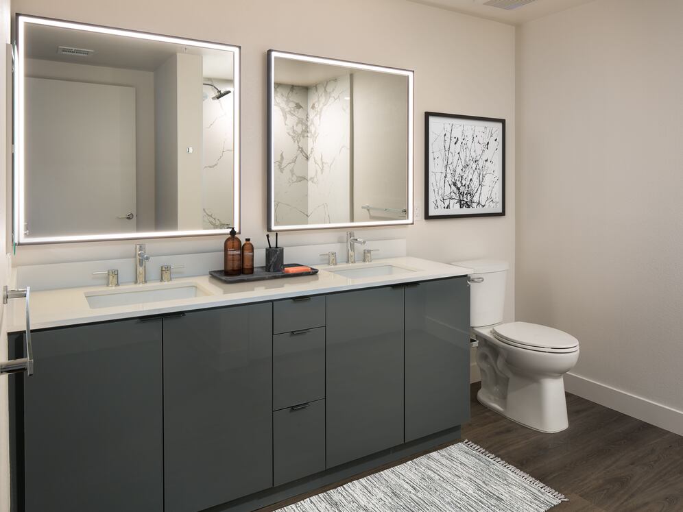 Upgrade Package Bath with grey cabinetry, white quartz countertop, marble shower surround, and lit vanity mirror