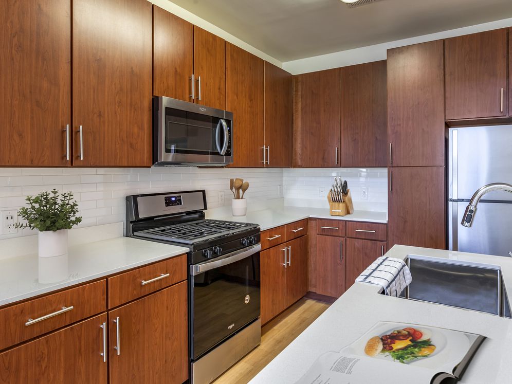 Newly renovated apartment homes feature kitchens with cherry cabinetry, white quartz countertops, stainless steel appliances, white tile backsplash, and hard surface flooring