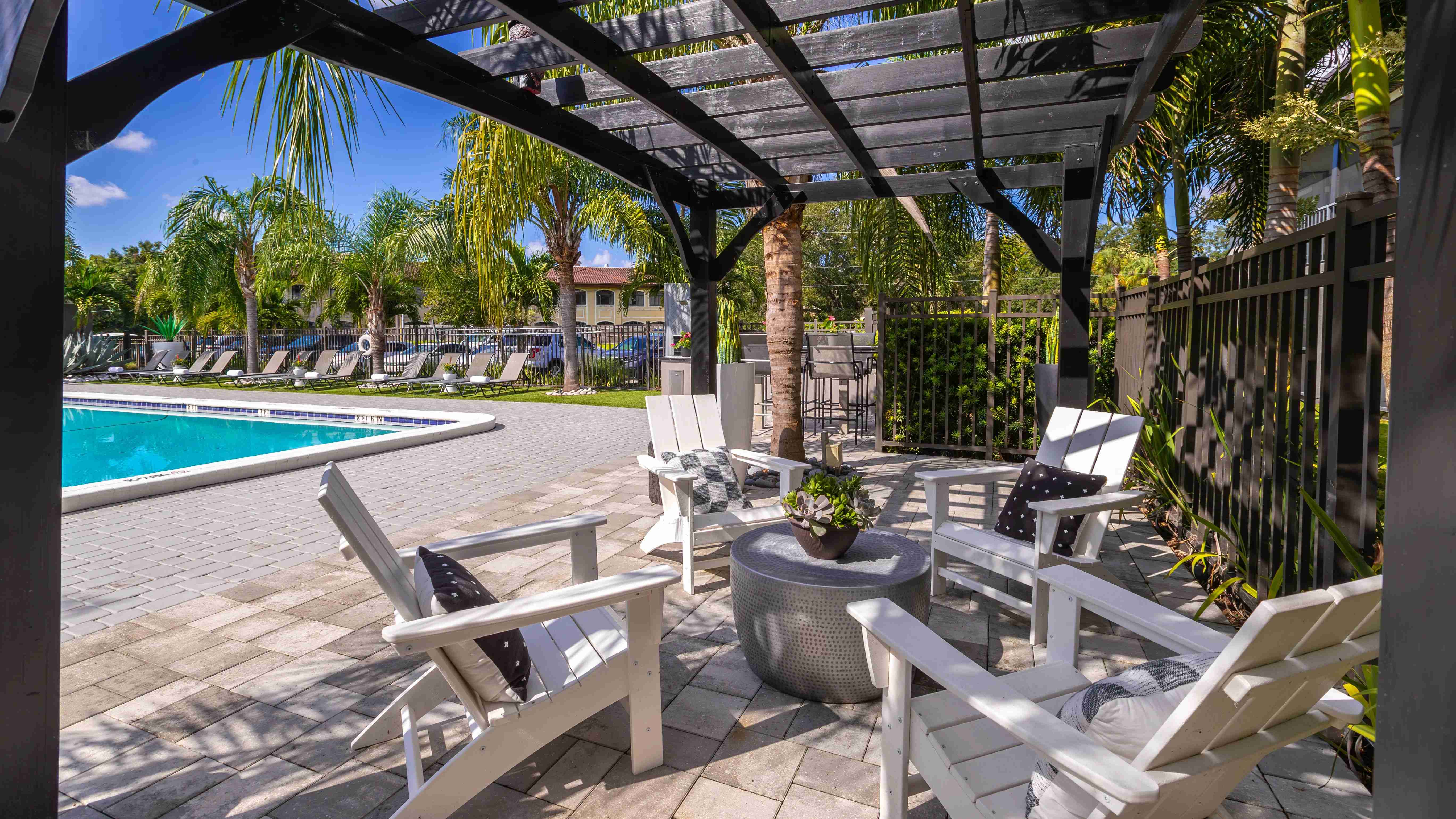 Relax in the comfortable, shaded poolside lounge featuring cozy seating under a pergola and views of the sparkling pool.